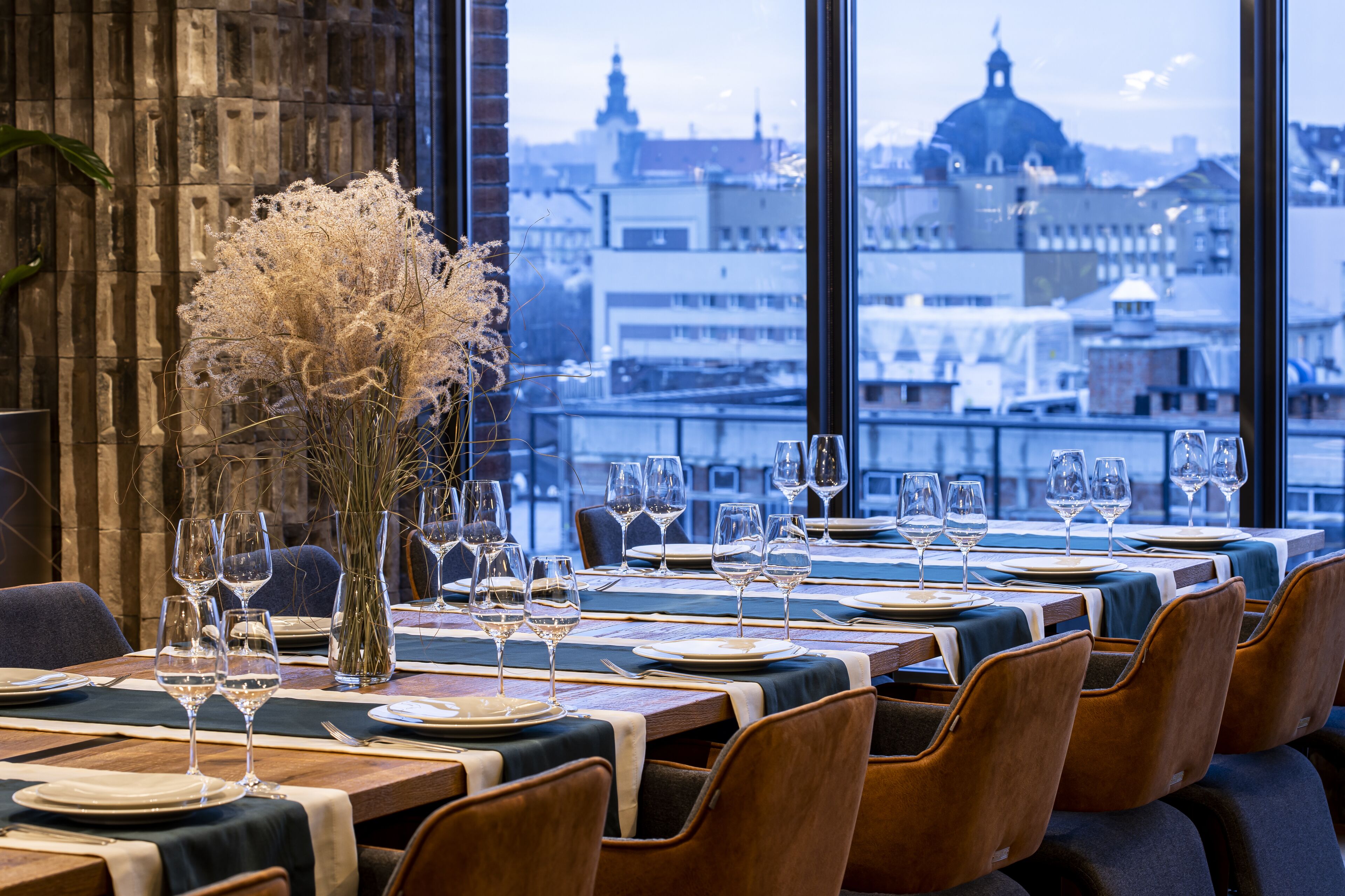 Un restaurant avec des tables dressées pour le dîner, un vase rempli de fleurs séchées et une vue sur le paysage urbain, visible à travers de grandes fenêtres.