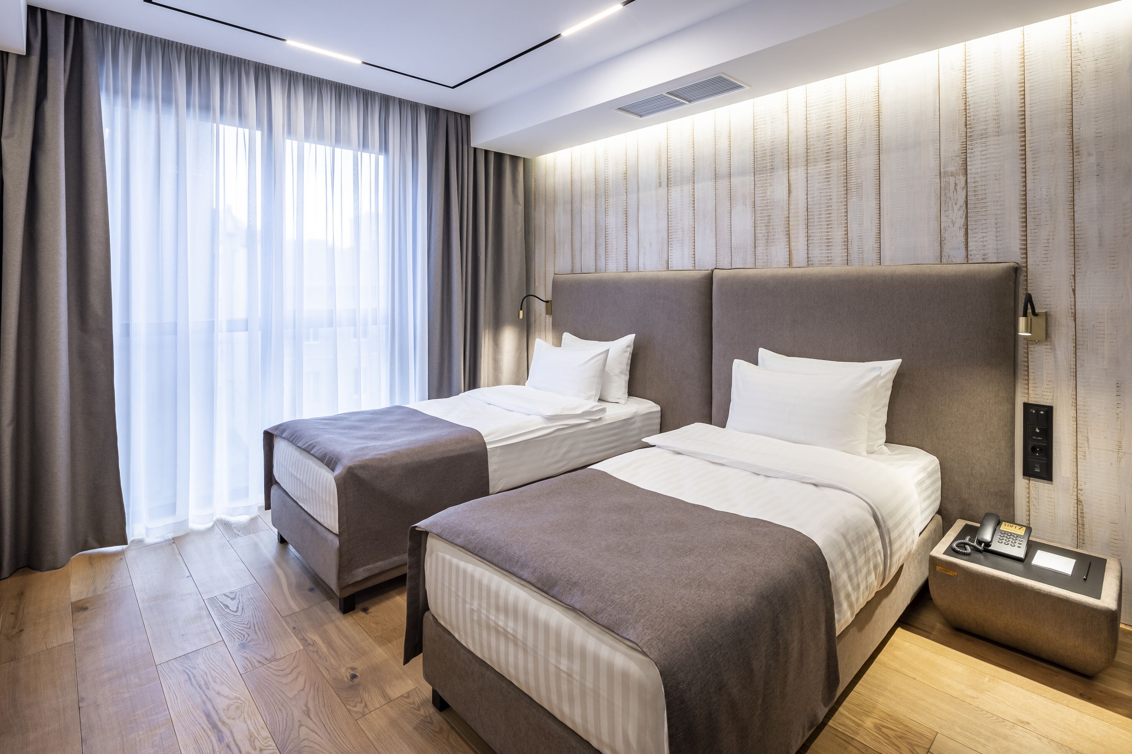 A hotel room with two beds, a bedside table with a phone, and a large window with curtains.