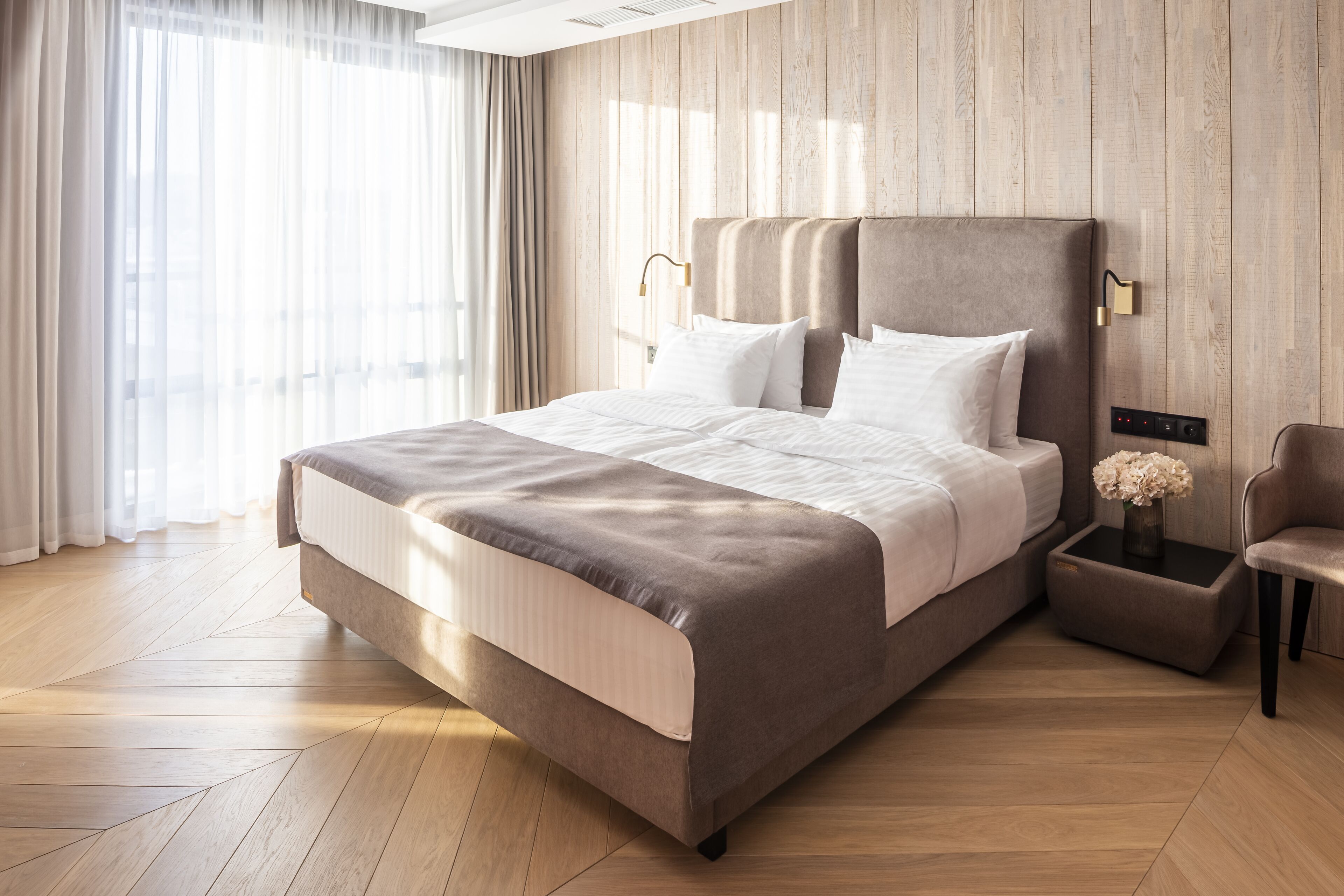 A modern hotel room with a large bed, a side table with a vase, and a chair.