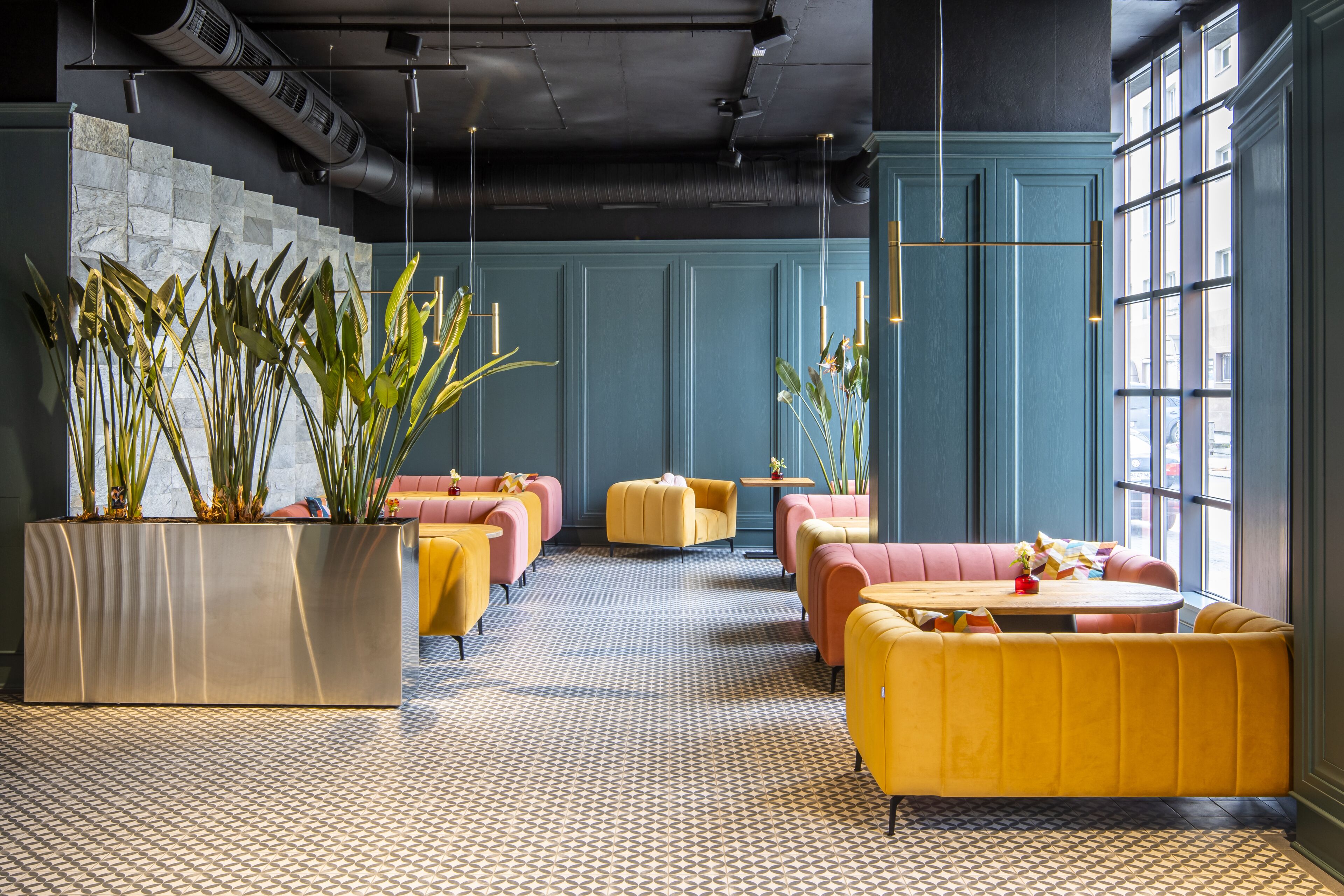 Eine moderne Lounge mit geometrischem Bodenmuster, gelb-rosa Sitzgelegenheiten und großen Topfpflanzen.