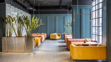 Eine moderne Lounge mit geometrischem Bodenmuster, gelb-rosa Sitzgelegenheiten und großen Topfpflanzen.