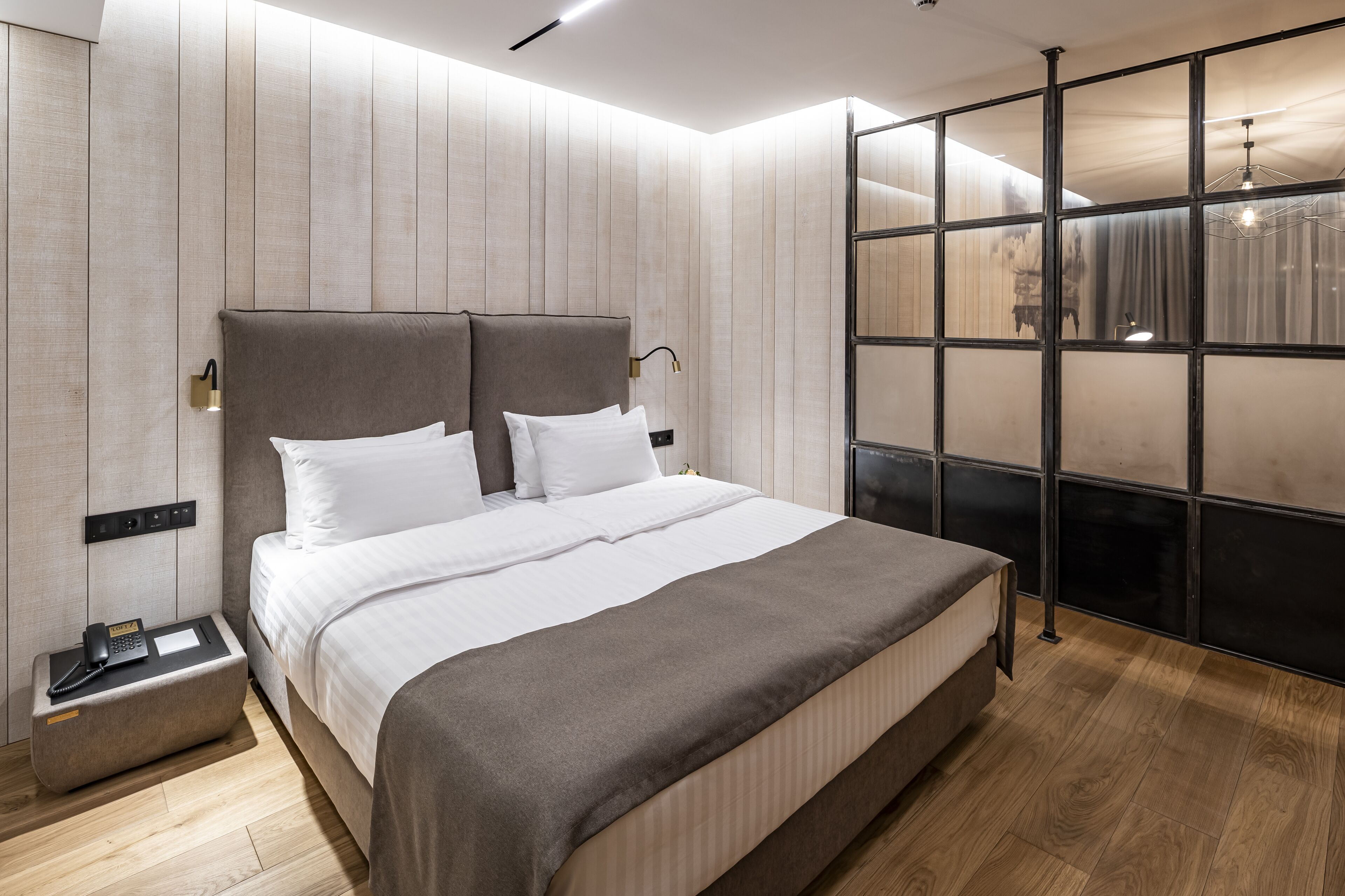 A modern hotel room with a large bed, a bedside table with a phone, and a view through a glass partition.