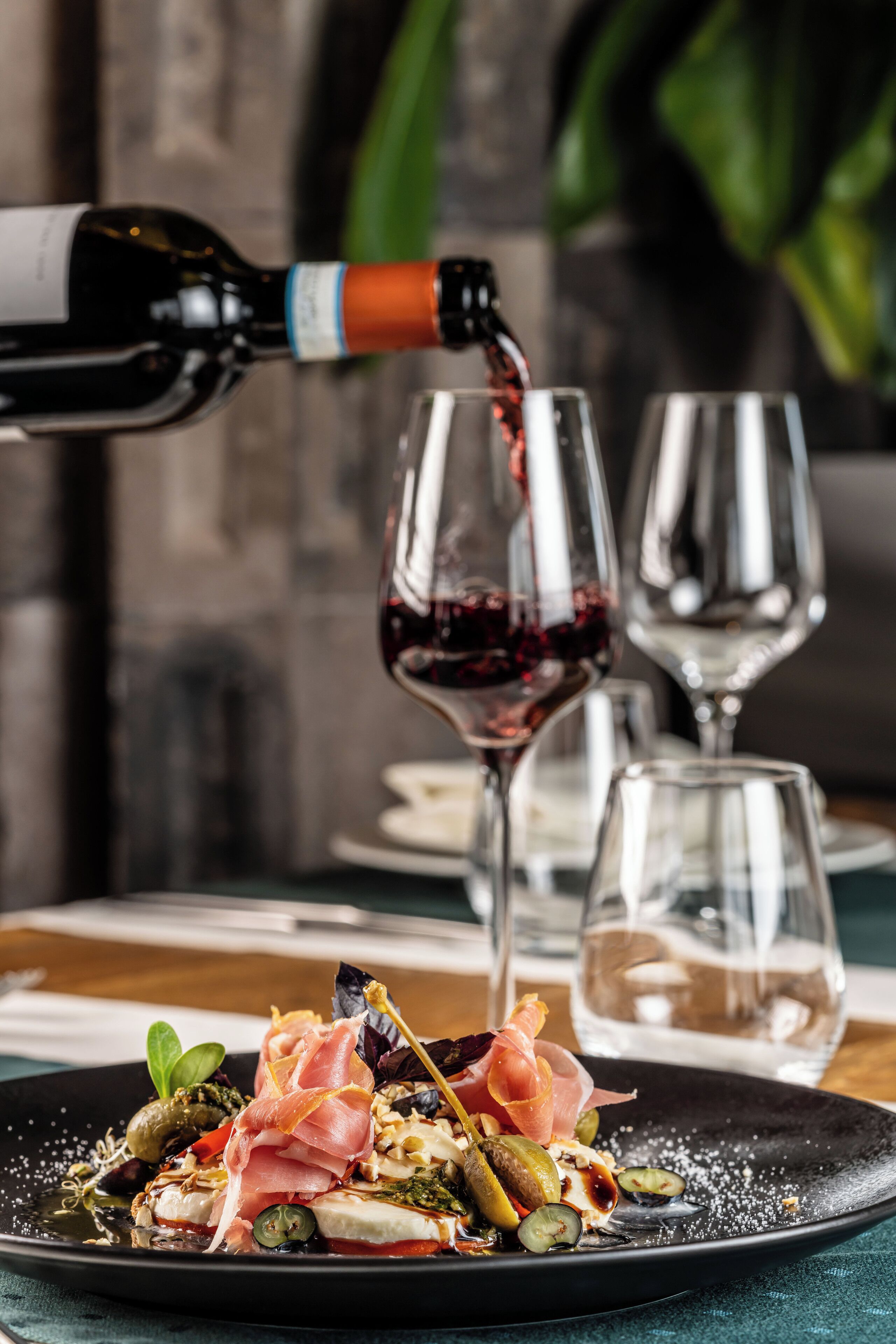 A dining table with a black plate of food, a wine bottle pouring into a glass, and two empty wine glasses.