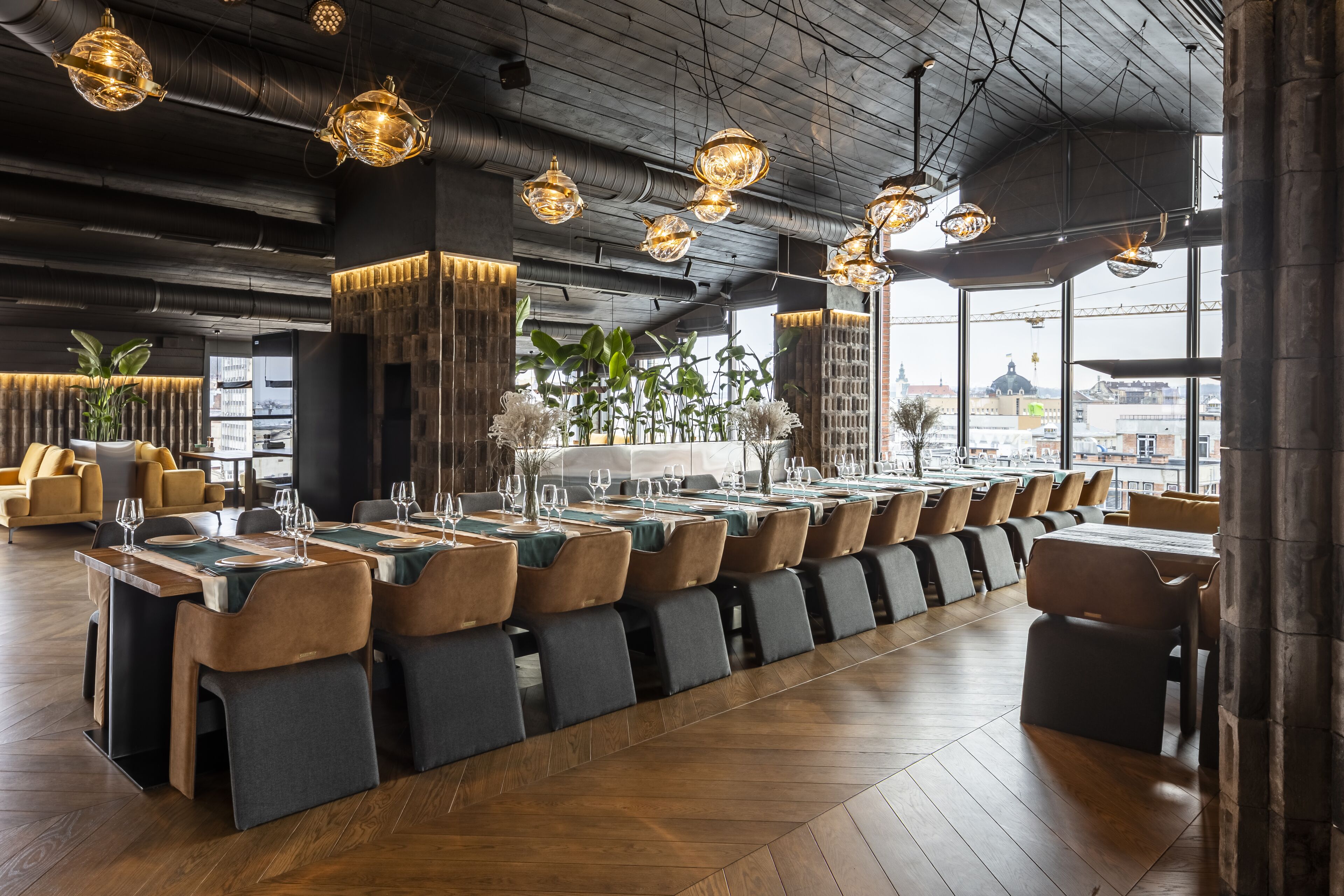 A modern restaurant with a long dining table, cushioned chairs, and hanging pendant lights.