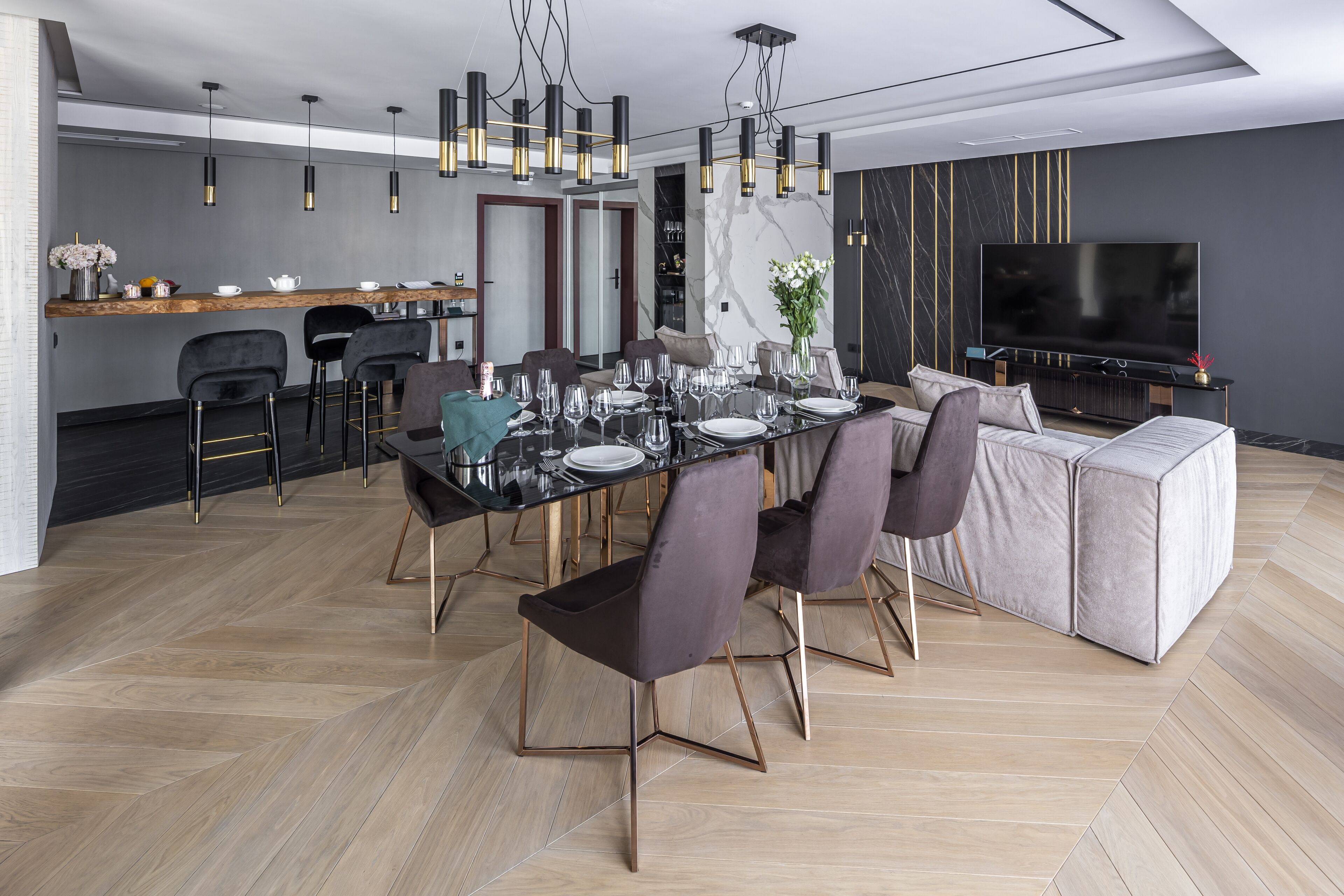Un espace de salle à manger moderne, avec une grande table dressée pour six personnes, entourée de chaises sombres aux pieds dorés. Un canapé et un téléviseur sont visibles au fond.
