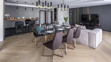 Un espace de salle à manger moderne, avec une grande table dressée pour six personnes, entourée de chaises sombres aux pieds dorés. Un canapé et un téléviseur sont visibles au fond.