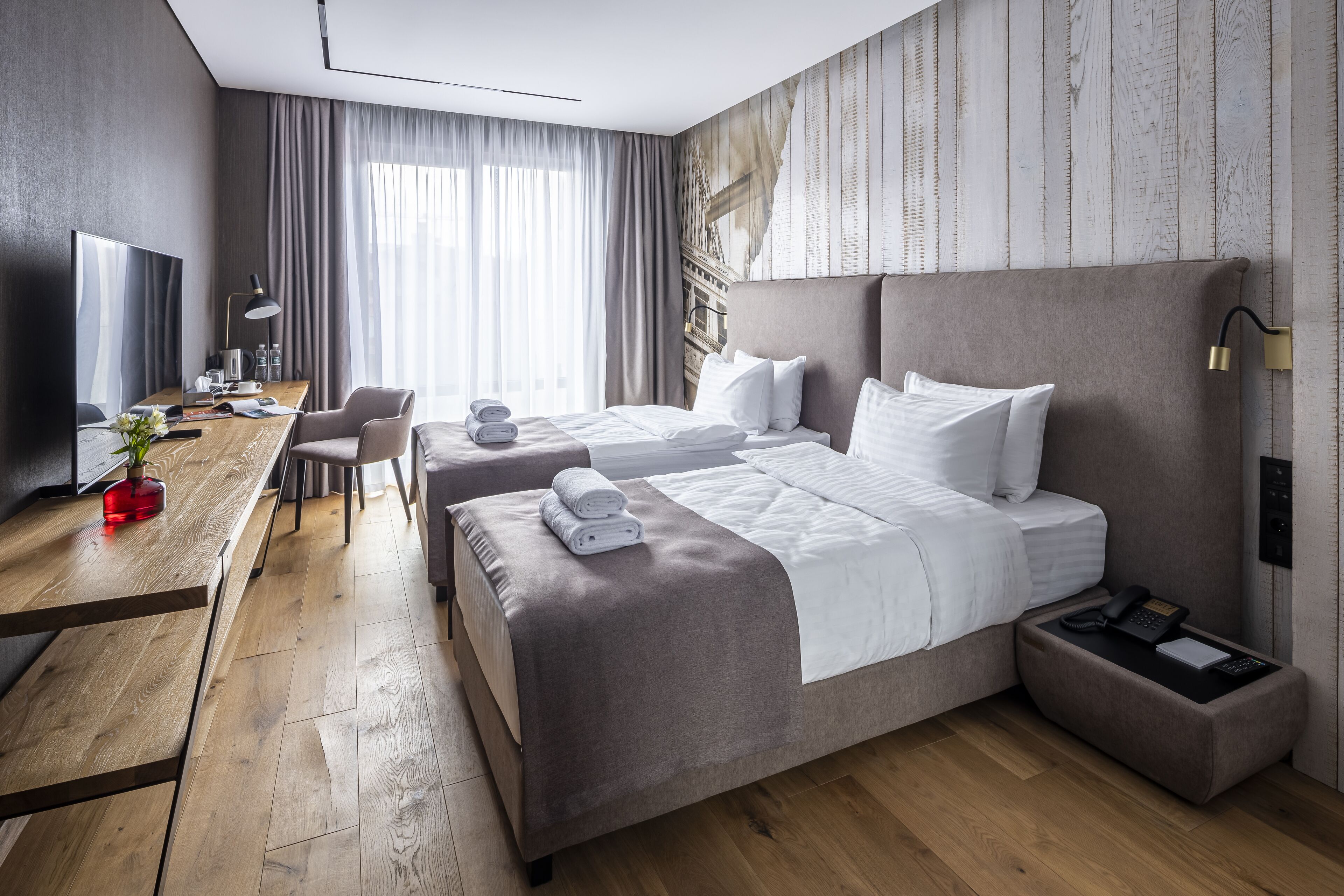 Moderni hotellihuone, jossa on kaksi sänkyä, työpöytä, televisio ja suuri ikkuna, jossa on verhot.