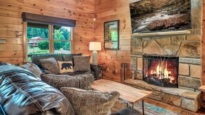 Ein gemütliches Blockhaus-Interieur mit Kamin aus Stein, Ledersofa und großem Fenster mit Blick auf einen Wald.
