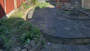 A residential backyard with a paved area, a wooden fence, and a brick house. - 5 Bedroom House (Upminster)
