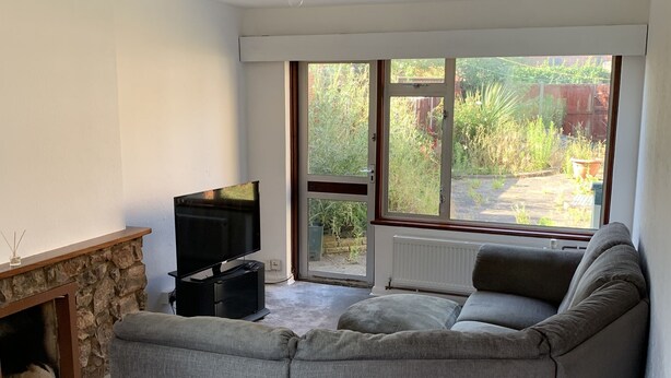 A living room with a stone fireplace, a grey sectional sofa, a flat-screen TV, and a large window overlooking a garden. - 5 Bedroom House (Upminster)