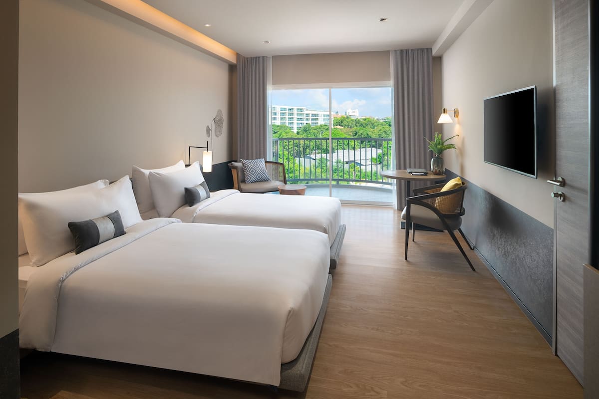 A hotel room with two beds, a desk, a chair, a TV, and a balcony with a view of buildings and trees.