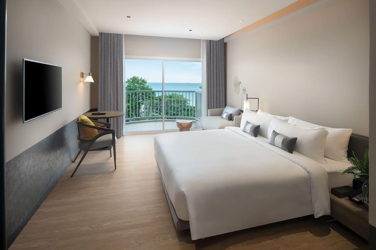 A modern hotel room with a large bed, a flat-screen TV, a small table with a chair, and a balcony with a view of the sea.
