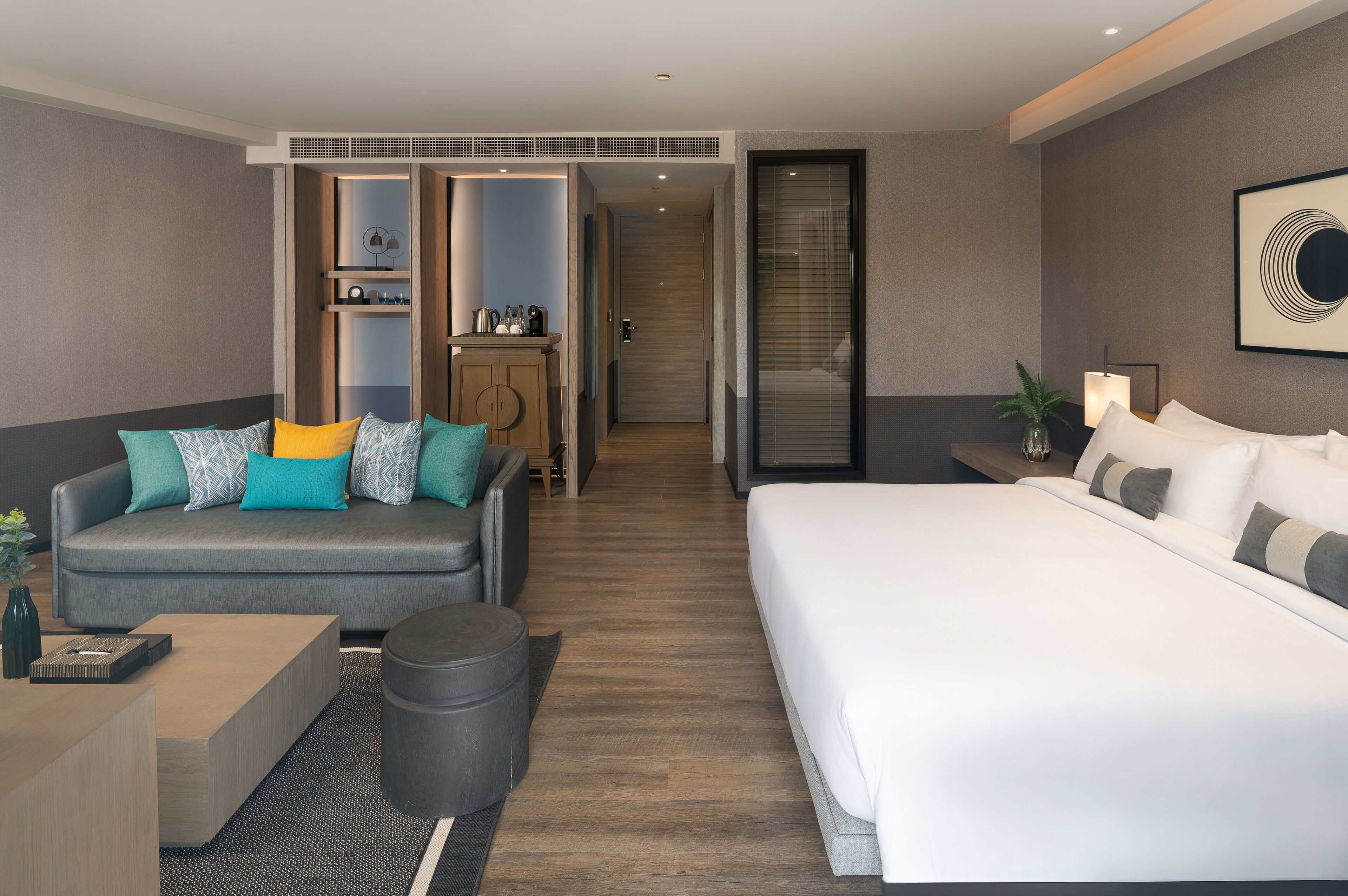 A modern hotel room with a grey sofa, a large bed, a wooden cabinet, and a small table.