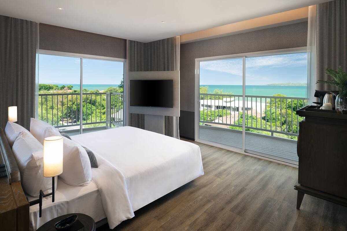 A hotel room with a large bed, a flat-screen TV mounted on the wall, and a balcony with a view of the ocean.