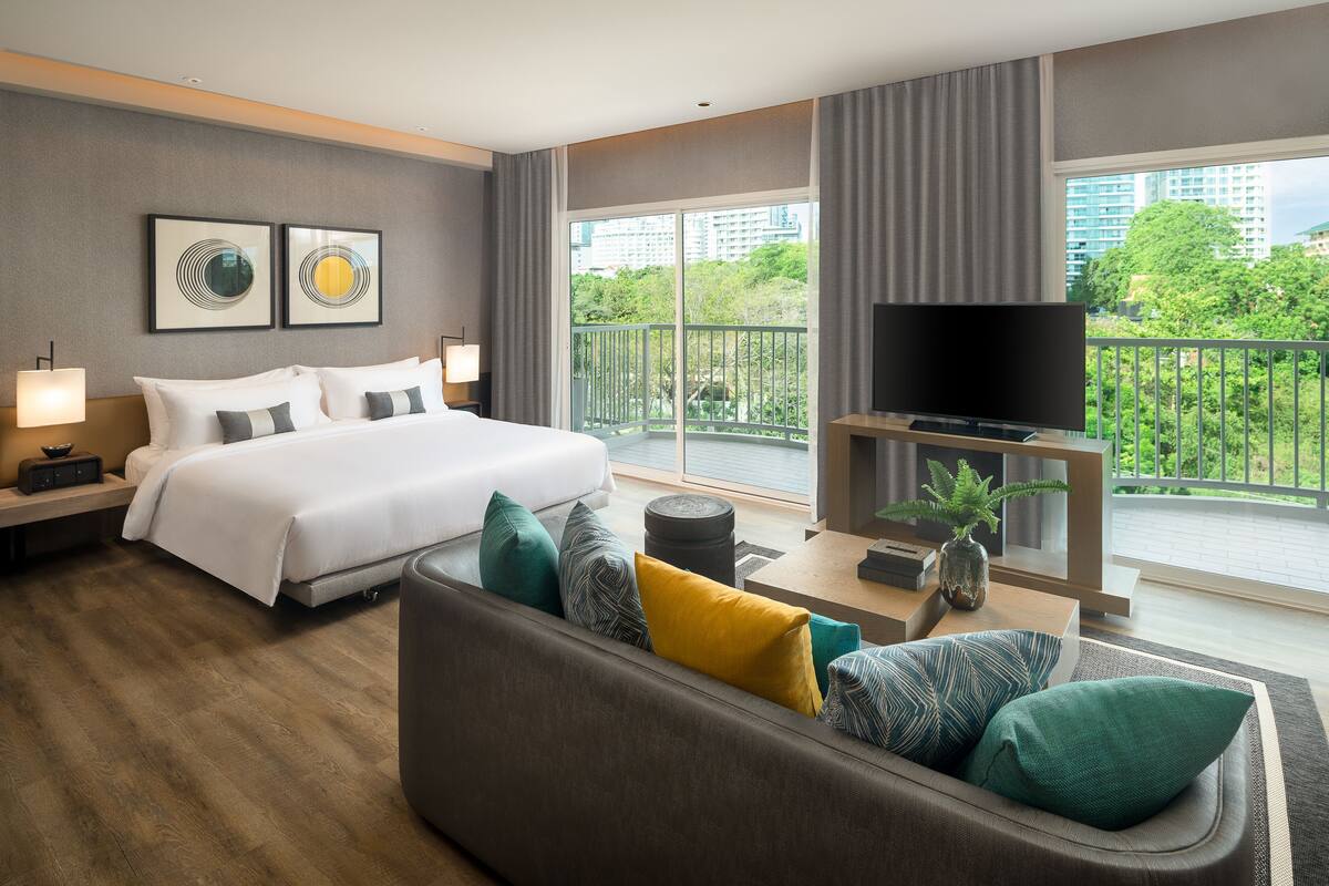 A hotel room with a bed, a sofa, a TV, and a balcony with a view of buildings and trees.