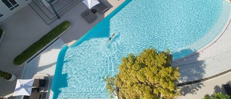 An aerial view of a swimming pool with lounge chairs and umbrellas, surrounded by a building with balconies and a tree.