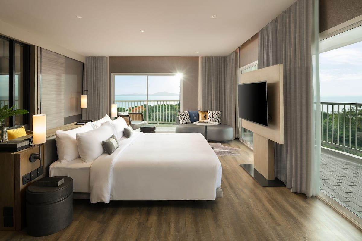 A modern hotel room with a large bed, a TV, a balcony with a view, and a small table with a lamp.