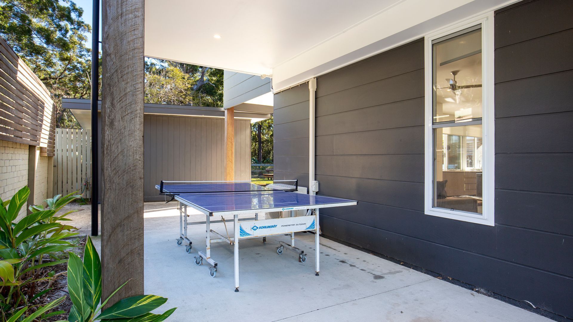 An outdoor ping pong table set up in a covered patio area with a view of the backyard and a house.