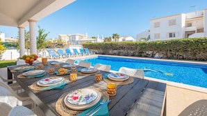 Un espace repas aménagé au bord de la piscine, avec une table dressée pour quatre personnes, donnant sur une piscine, entouré de chaises longues et d’une haie.
