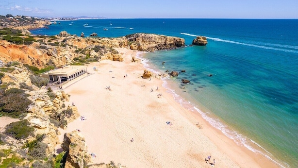 Une plage côtière avec un terrain rocheux, une zone de plage sablonneuse et un bâtiment doté d’un toit en pente, situé près de l’eau.