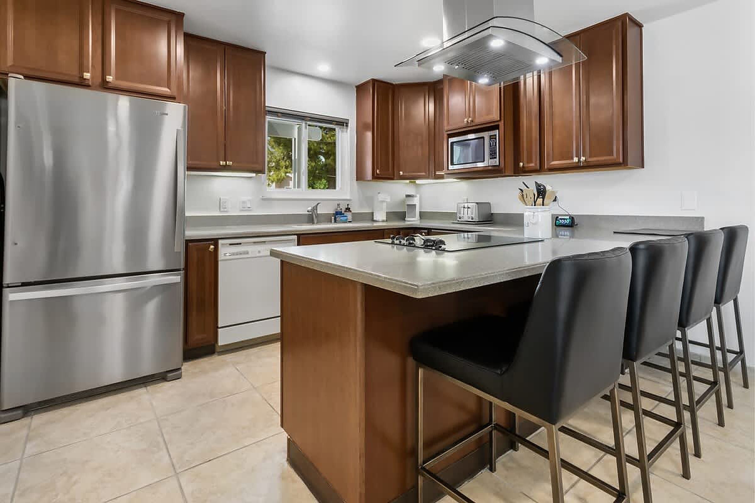 Una cocina moderna con electrodomésticos de acero inoxidable, armarios de madera, una isla central con taburetes y un ventanal que proporciona luz natural.