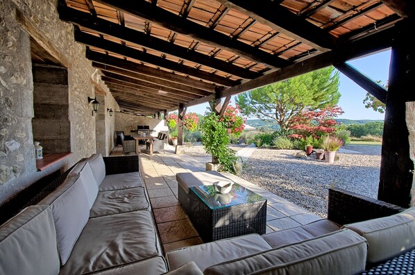 An outdoor patio with a stone wall, wicker furniture, a glass-top table, and a view of trees and hills.