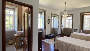 Un dormitorio espacioso con una cama grande, un aparador y un tocador. Varias ventanas dejan entrar luz natural y un candelabro cuelga del techo.