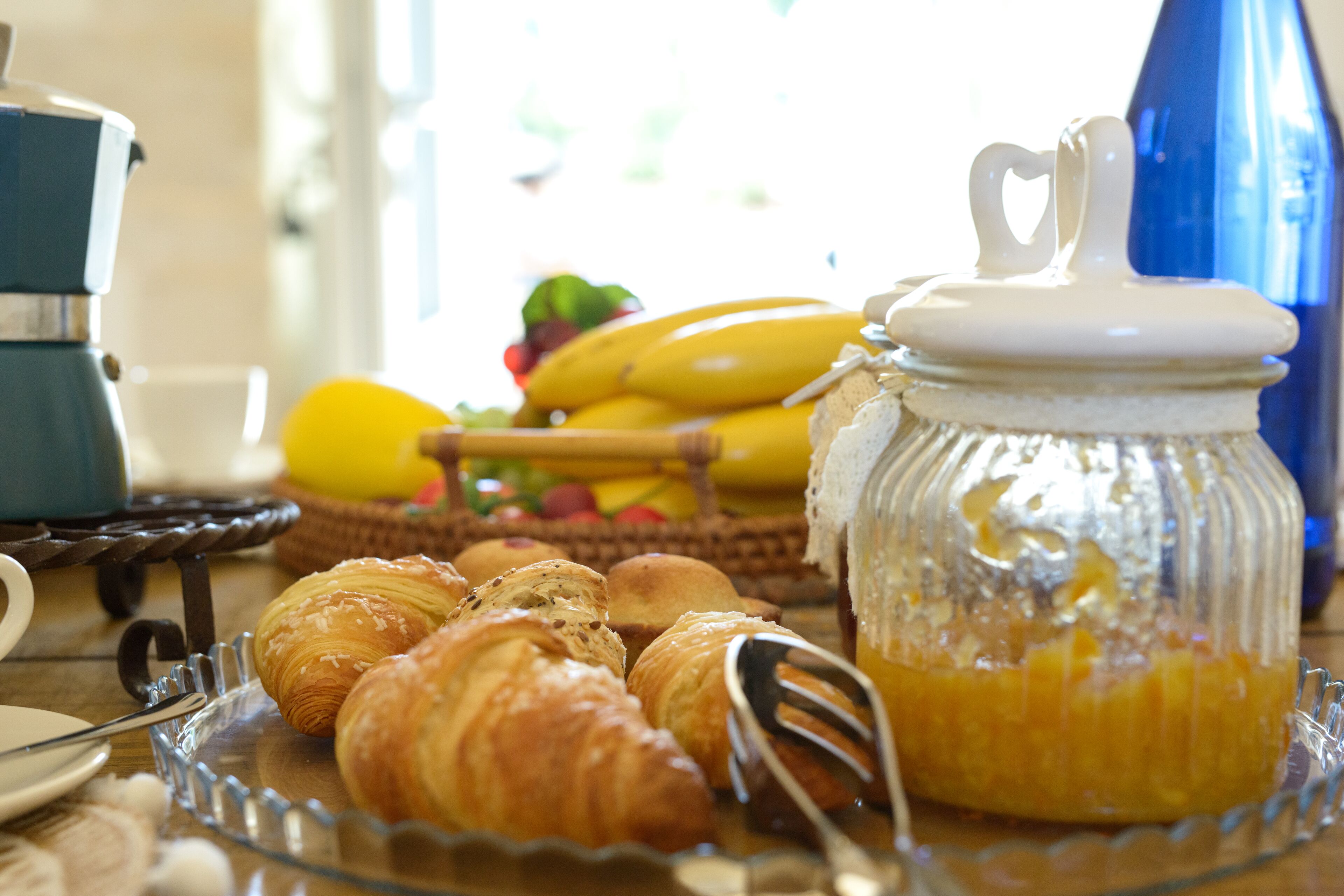 Una escena de desayuno con cruasanes, un frasco de mermelada y una cesta de fruta.