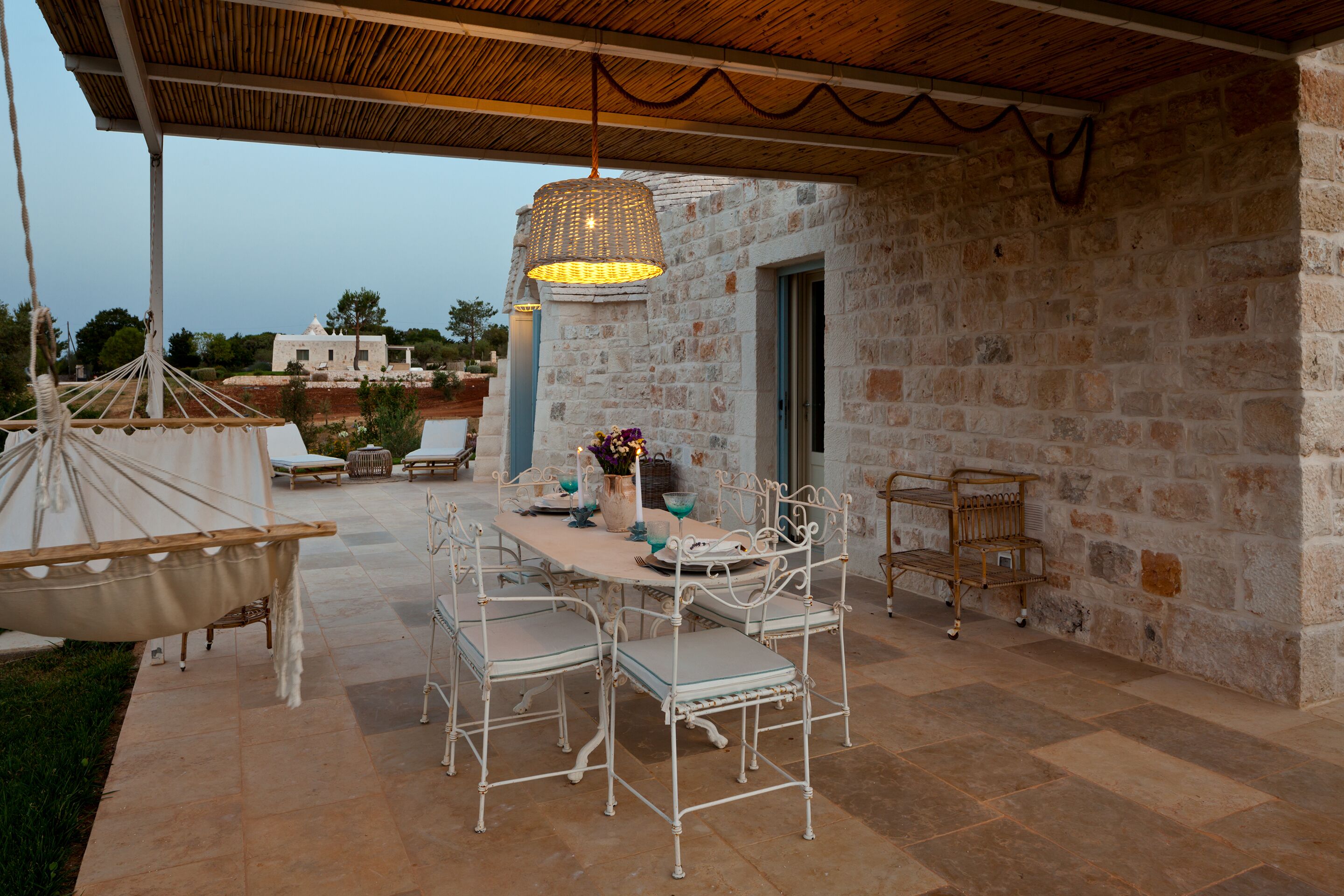 Une terrasse extérieure avec un mur en pierre, un luminaire suspendu, une table dressée pour quatre personnes et un hamac.