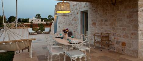 Une terrasse extérieure avec un mur en pierre, un luminaire suspendu, une table dressée pour quatre personnes et un hamac.