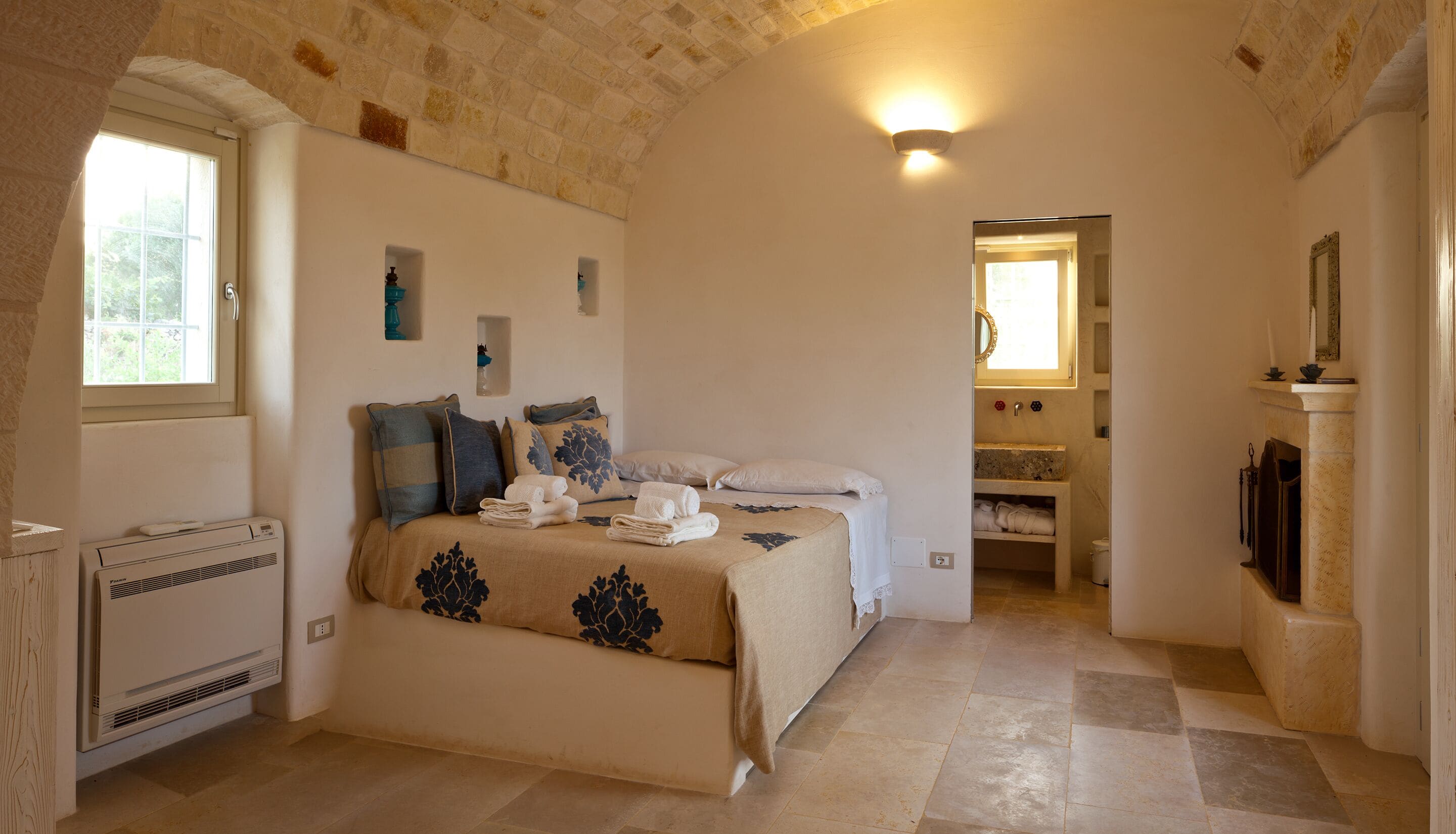 Een slaapkamer met een bed, een radiator, een raam en een badkamer met een spiegel en wastafel.