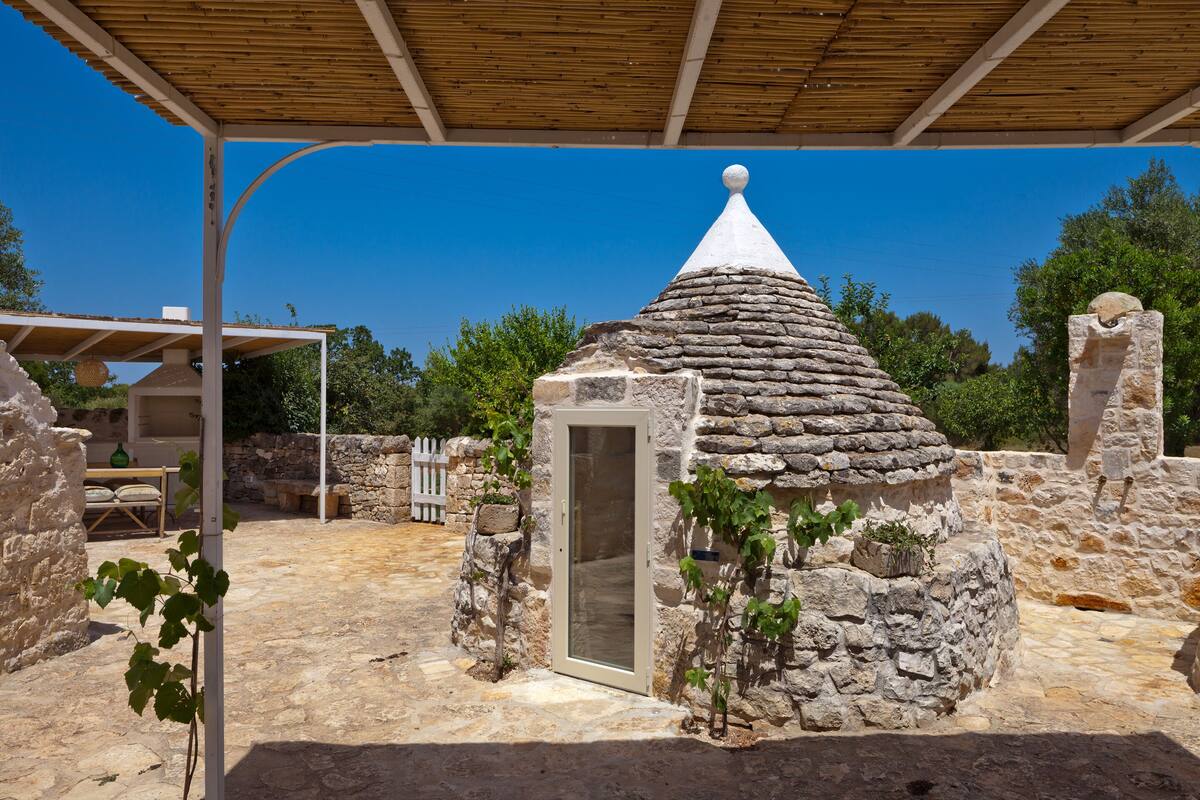 A stone-built structure with a conical roof and a door, surrounded by stone walls and greenery.