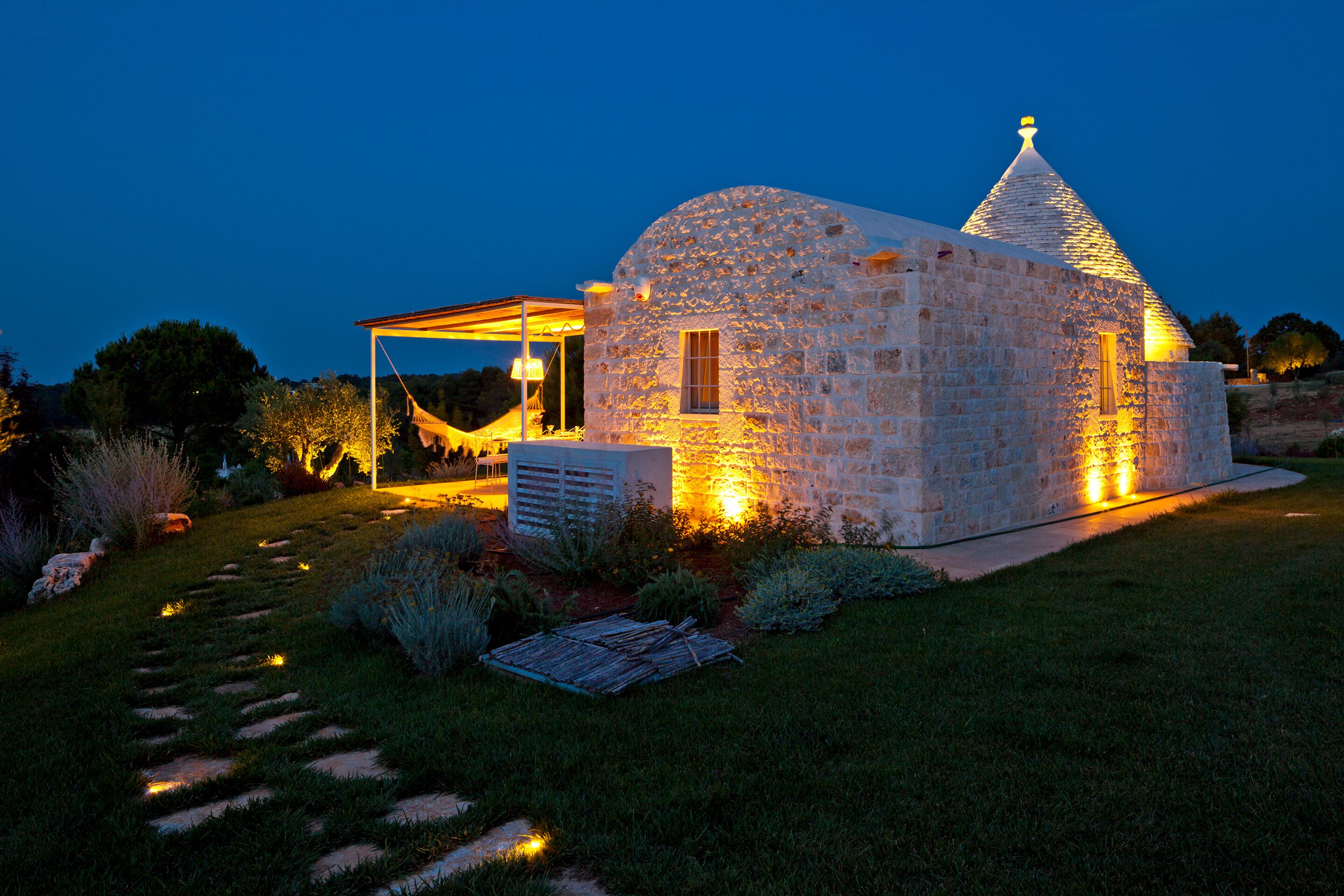 A stone house with a conical roof, illuminated by warm lighting, surrounded by a well-maintained garden with a stone pathway.