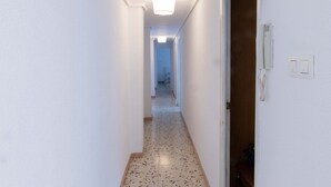 A narrow hallway with white walls and a speckled floor, leading to a room with a wooden door and a light switch on the wall.