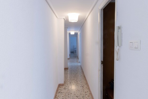 Een smalle gang met witte muren en een gespikkelde vloer, die leidt naar een kamer met een houten deur en een lichtschakelaar aan de muur.
