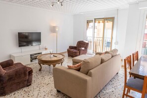 Een woonkamer met een bank, fauteuils, een televisie, een salontafel en een eettafel met stoelen.
