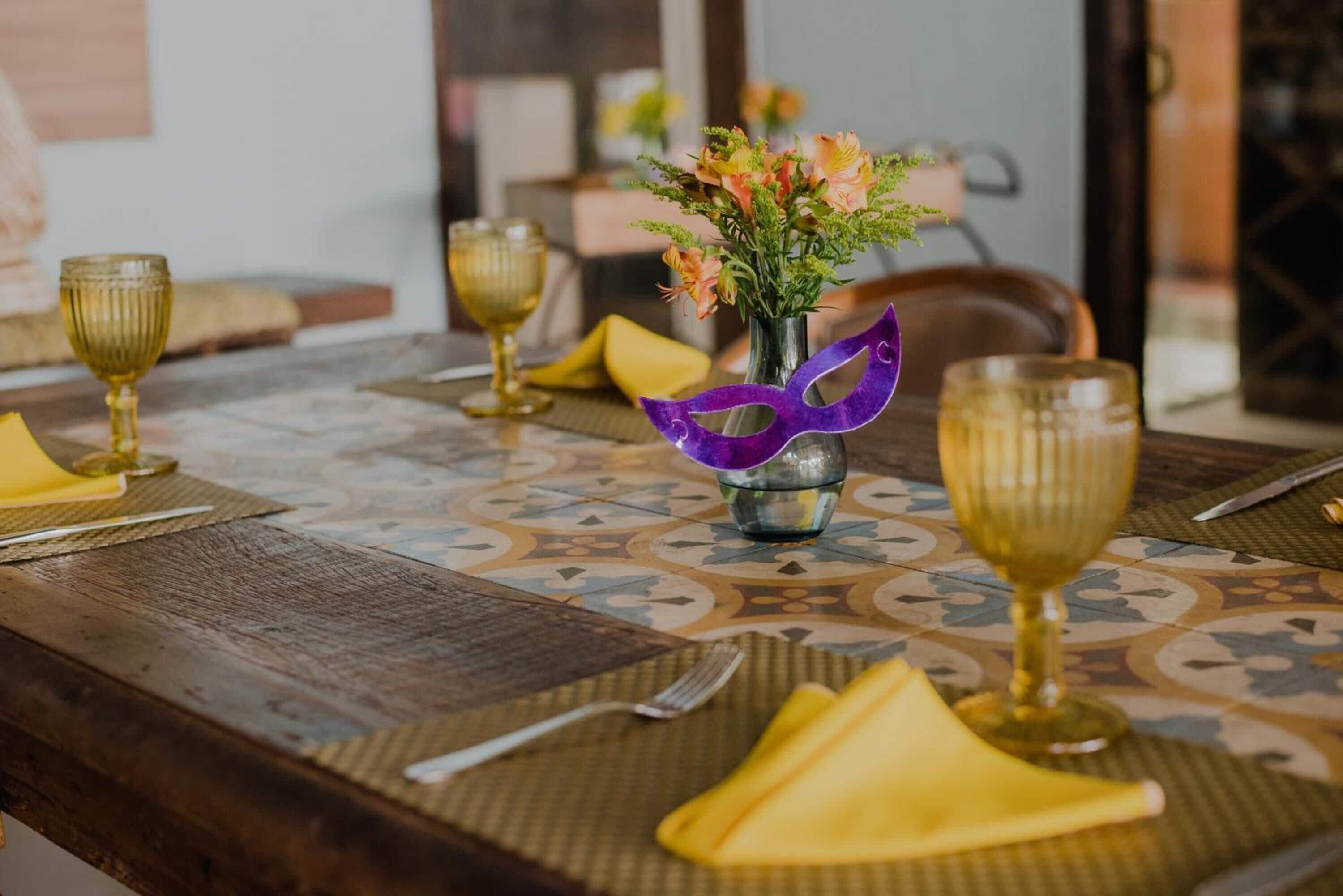 A dining table set with yellow napkins, a vase with flowers, and a purple mask.