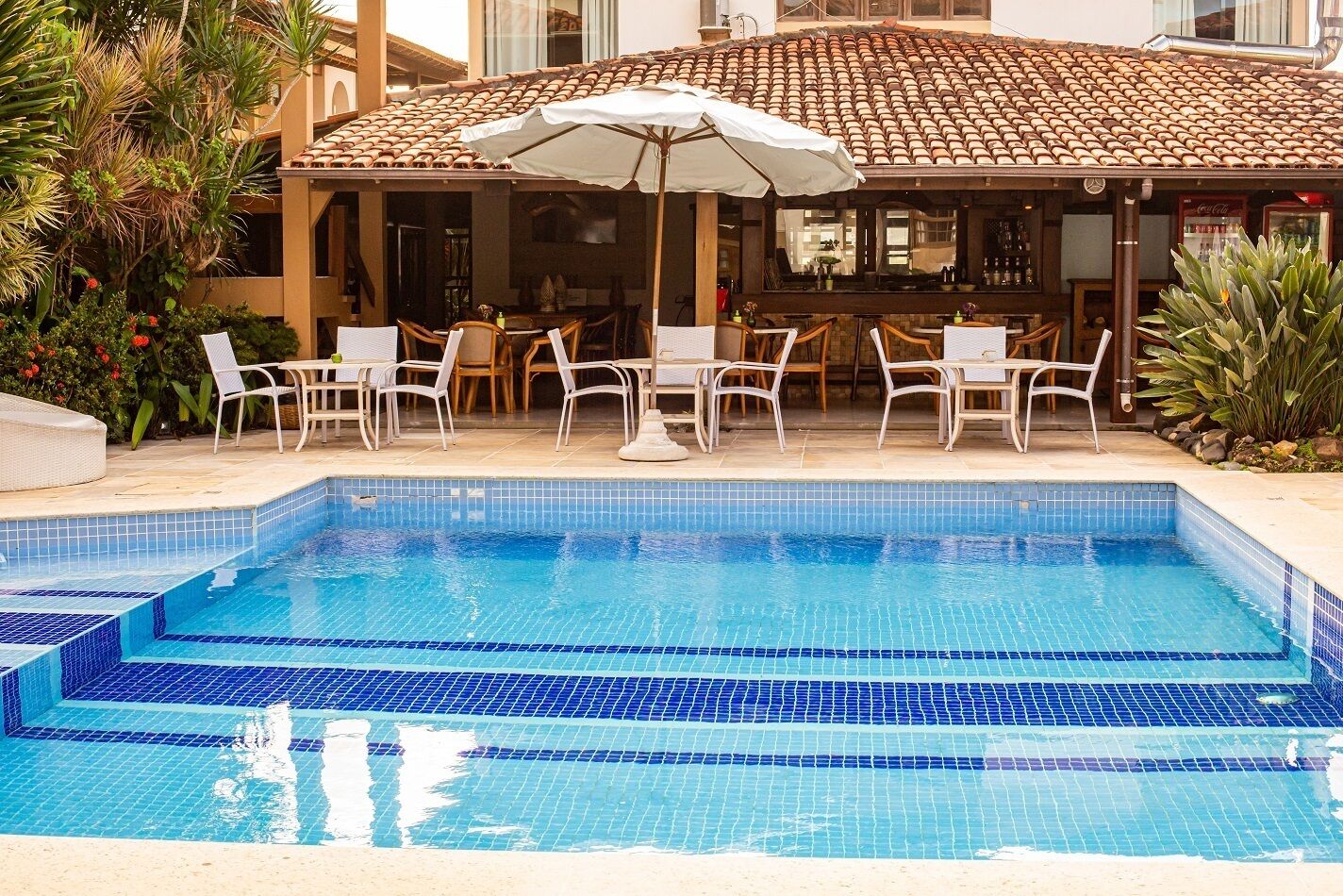 Piscina rectangular con baldosas azules, rodeada de sillones blancos y una sombrilla.
