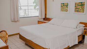 A clean, well-lit bedroom with a wooden bed, a small bedside table, a chair, and a window with curtains.