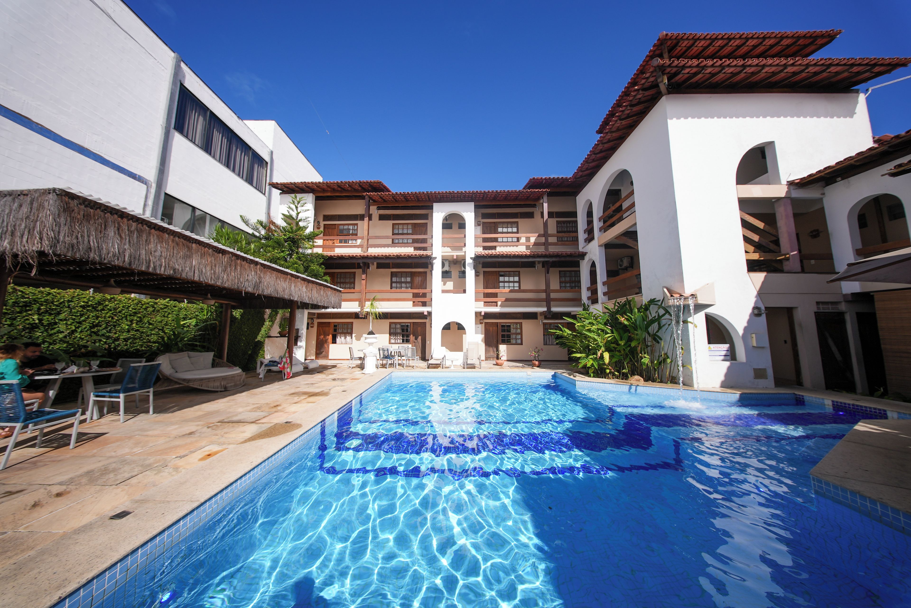 A swimming pool with a clear blue water surface, surrounded by a building with multiple balconies and a thatched roof structure.