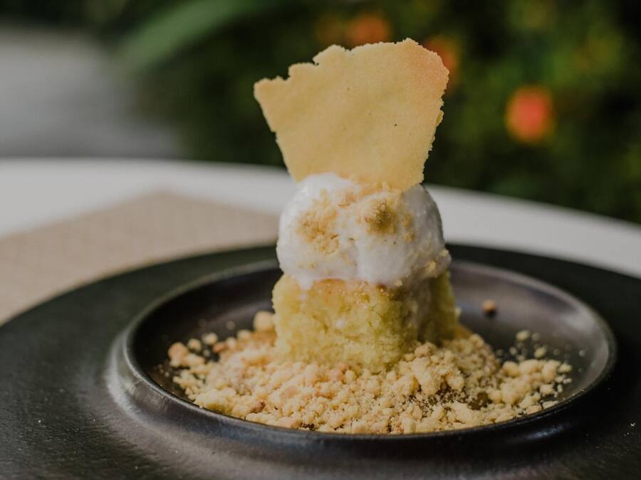 Un postre con una porción de helado encima de un pastel, servido en un plato negro rodeado de migas.