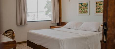Quarto com cama de madeira, janela com cortinas, uma cadeira e duas obras de arte penduradas na parede.