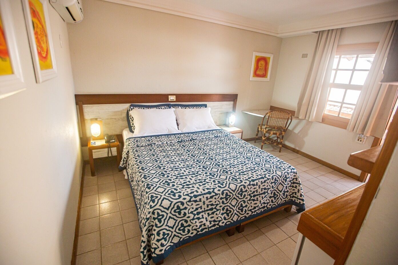 A hotel room with a large bed, a bedside table with a lamp, a chair, and a window with curtains.