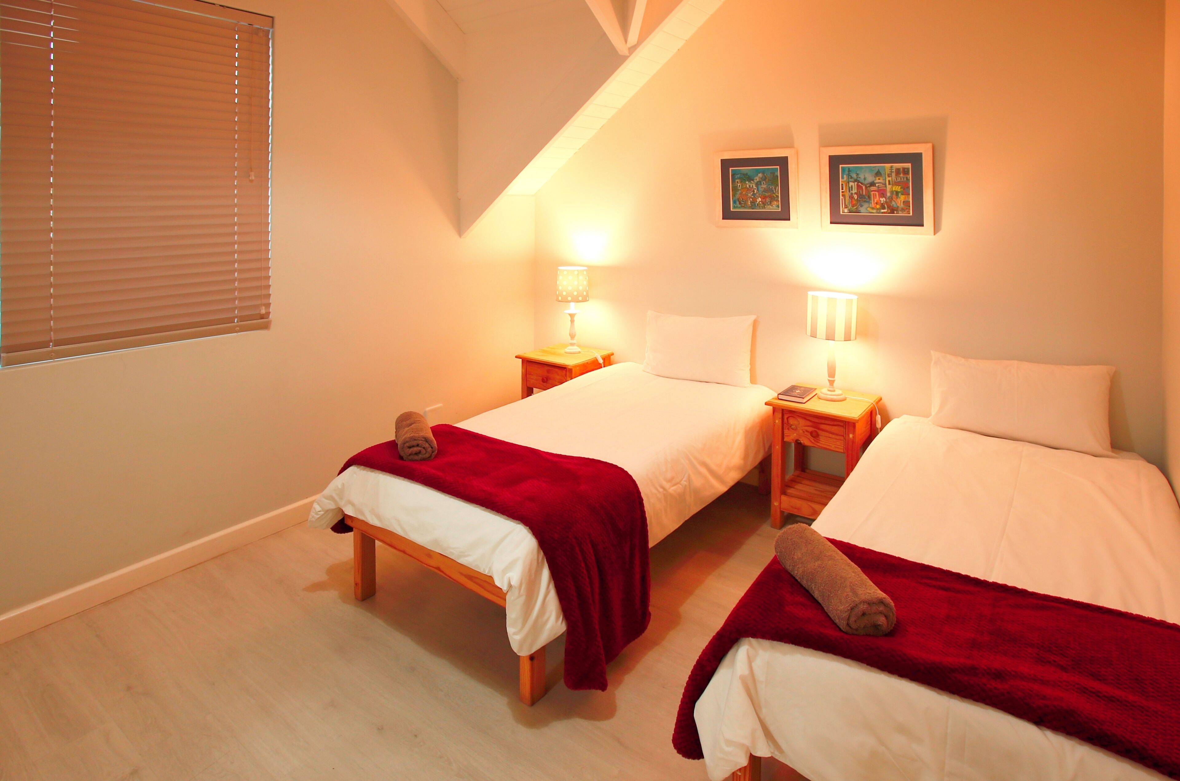 A bedroom with two single beds, each with a red blanket and a rolled towel. There are bedside tables with lamps, and two framed pictures on the wall.