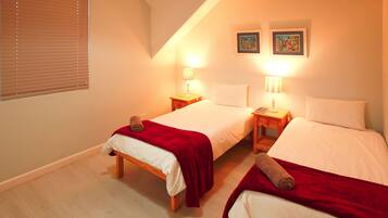 A bedroom with two single beds, each with a red blanket and a rolled towel. There are bedside tables with lamps, and two framed pictures on the wall.