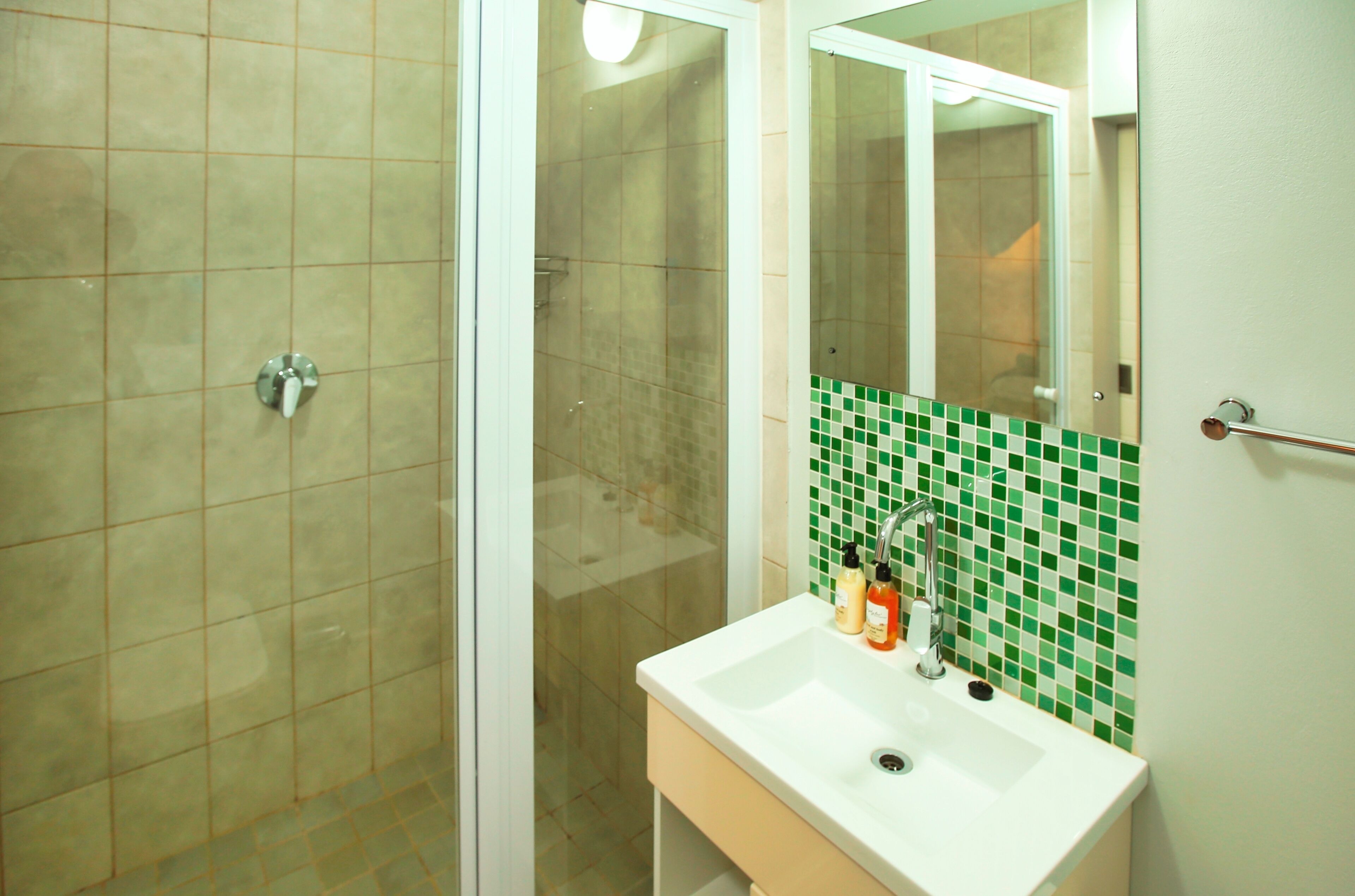 Een badkamer met een glazen douchedeur, een wit wastafelmeubel en een spiegel met een groene mozaïektegel achterwand.
