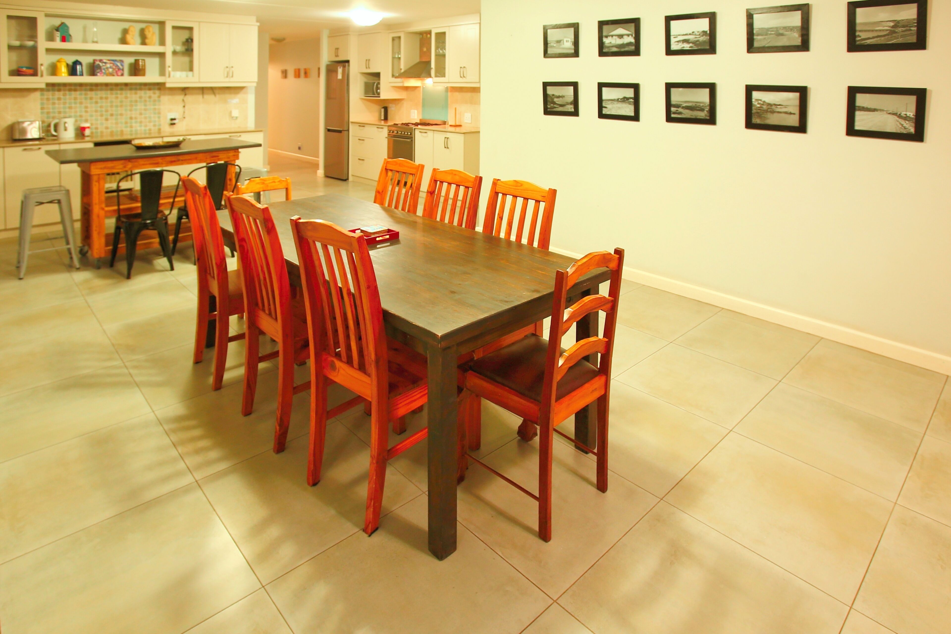 A dining area with a wooden table and six chairs, a kitchen in the background, and a wall with framed pictures.