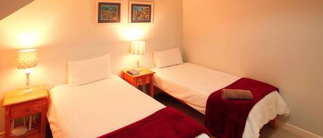 A room with two single beds, each with a red blanket and pillow, a nightstand with a lamp, and two framed pictures on the wall.