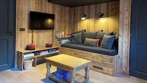 Un salon chaleureux avec un mur en bois, un téléviseur à écran plat fixé au-dessus d’un canapé agrémenté de coussins, une petite table en bois et une bibliothèque remplie de livres et de jeux de société.