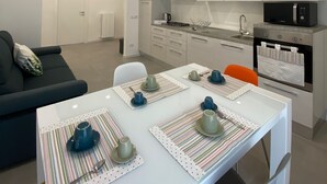 Cocina moderna con armarios blancos, un microondas negro y un horno. Hay una mesa de comedor con individuales a rayas y tazas azules. Un sofá negro y una silla blanca están en la sala de estar.